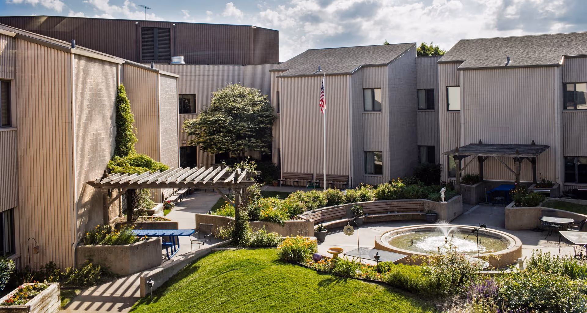 Sunny courtyard with a circular fountain, benches, pergola, flagpole and surrounding multi-story residential buildings.