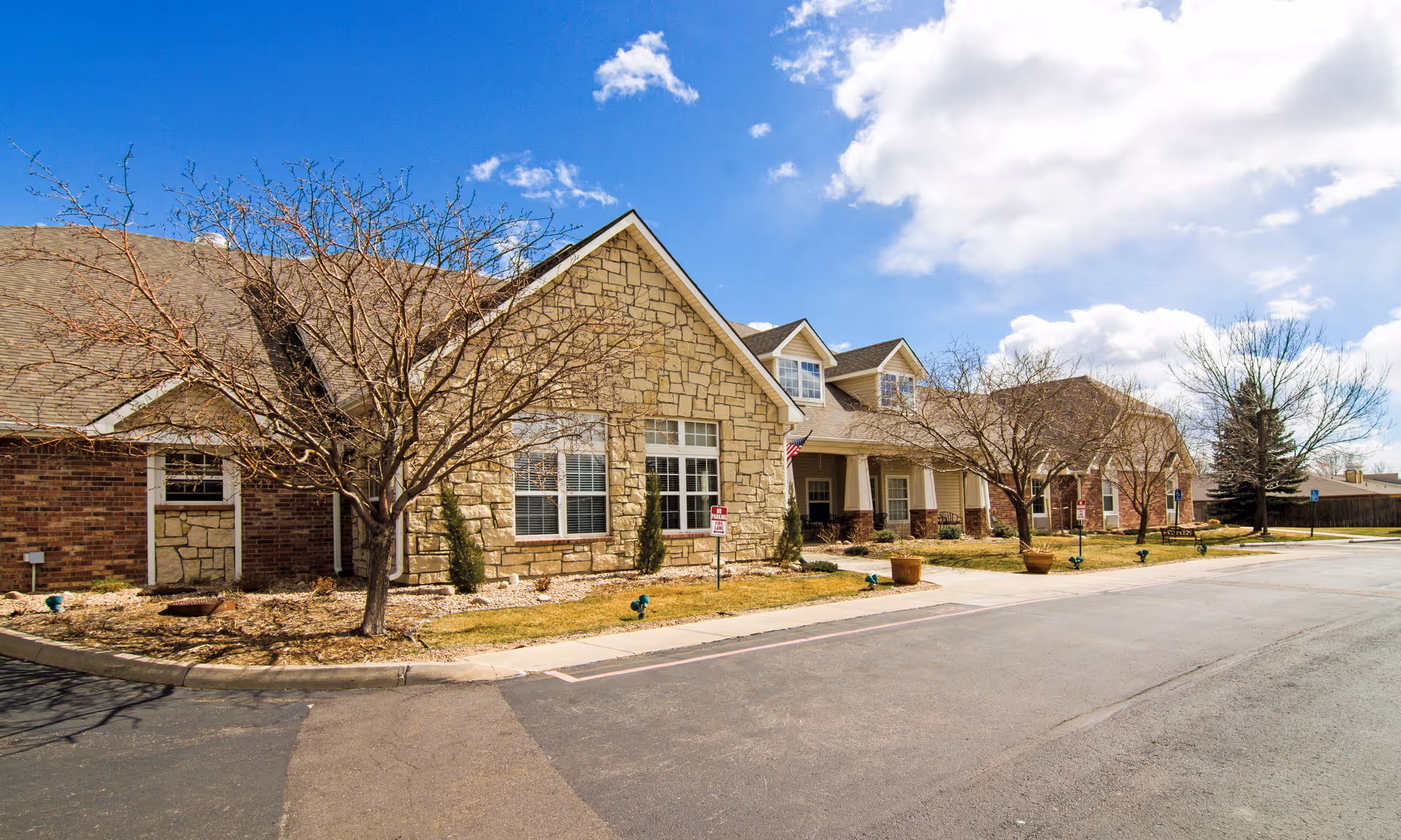 Stone-facade senior living facility exterior with peaked roof, leafless trees, and a curbside street.