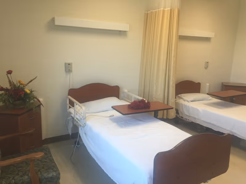 Two hospital-style beds in a shared patient room with overbed tables, a curtain divider, and a flower arrangement on a bedside cabinet.