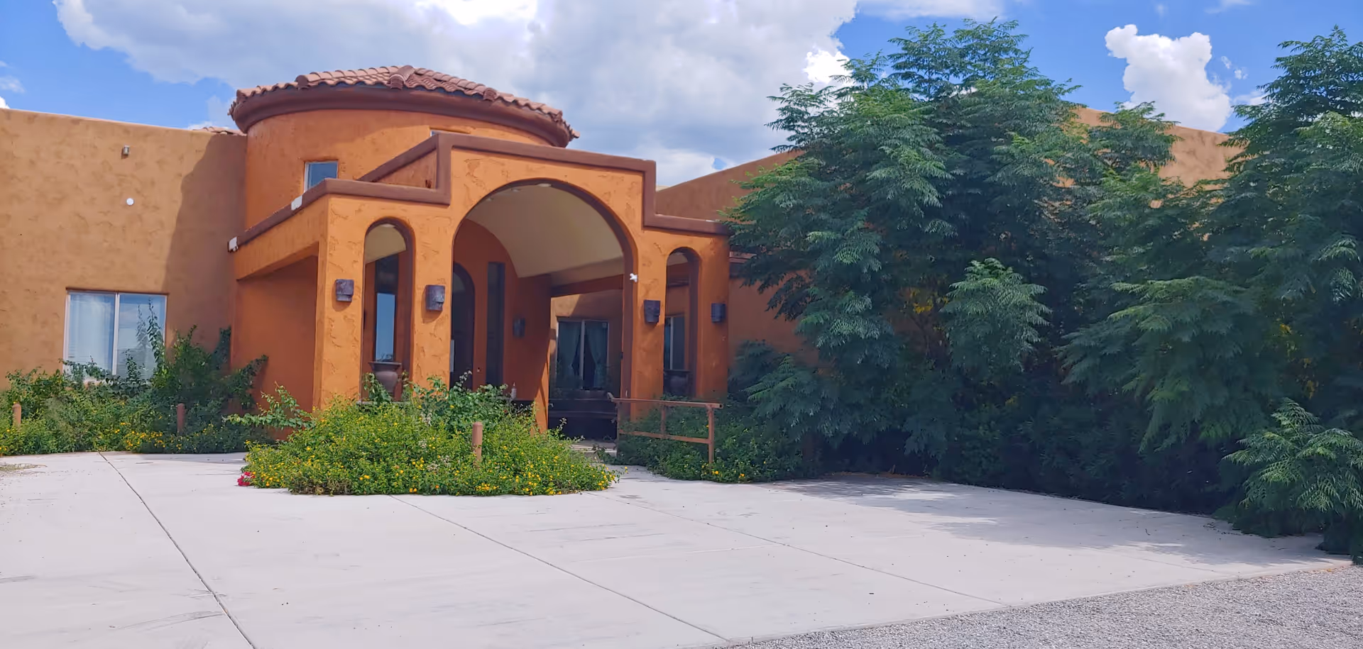 Exterior view of a building with a southwestern architectural style featuring an arched entrance, terracotta-colored walls, and a tiled roof. The entrance is surrounded by greenery and bushes, with a concrete driveway in front. The sky is partly cloudy.