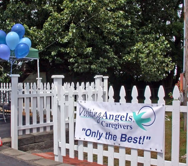 White picket fence with a banner reading Visiting Angels Caregivers Only the Best and blue balloons.