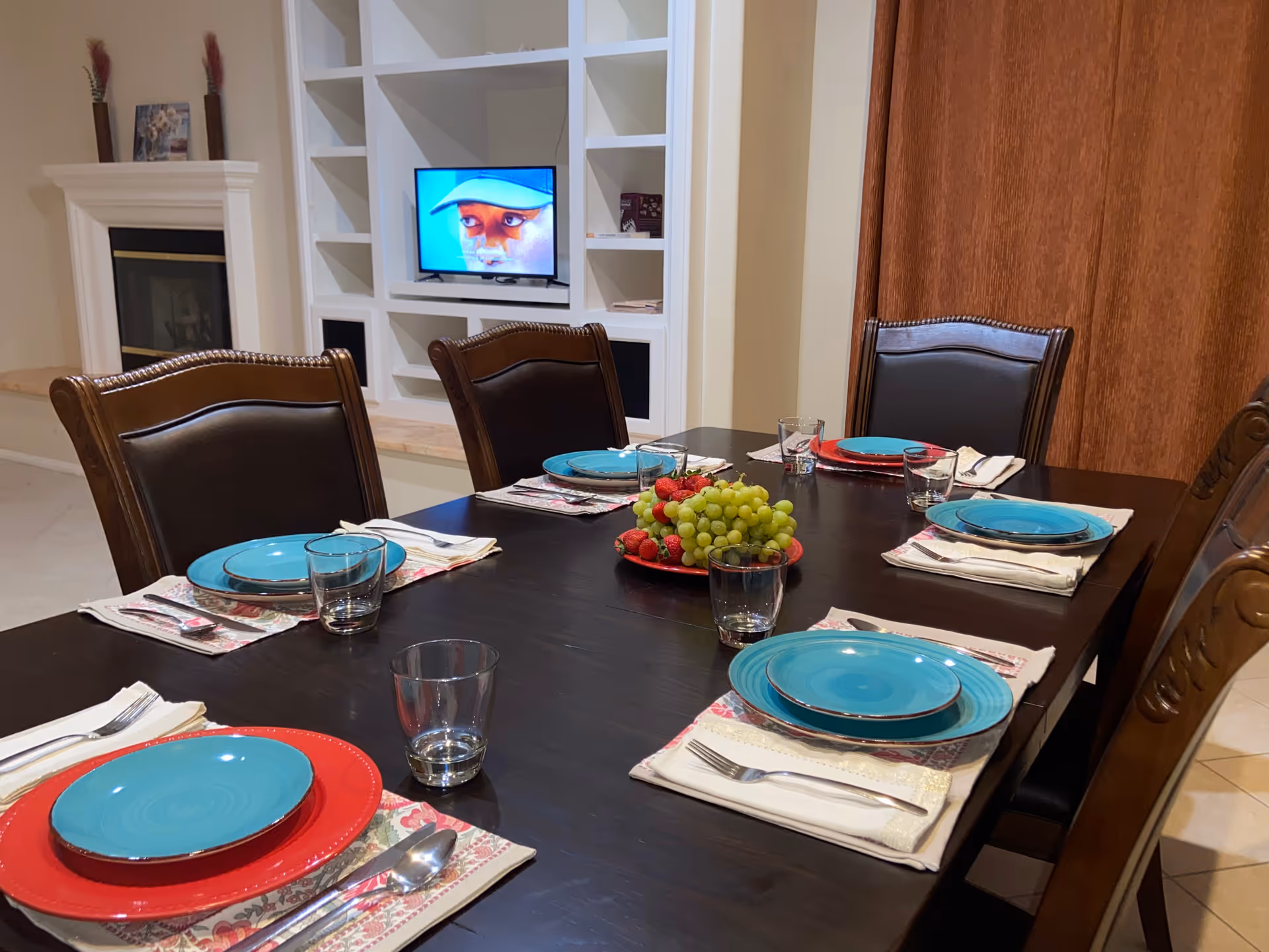 A dining room table set for a meal with a TV and shelves in the background.