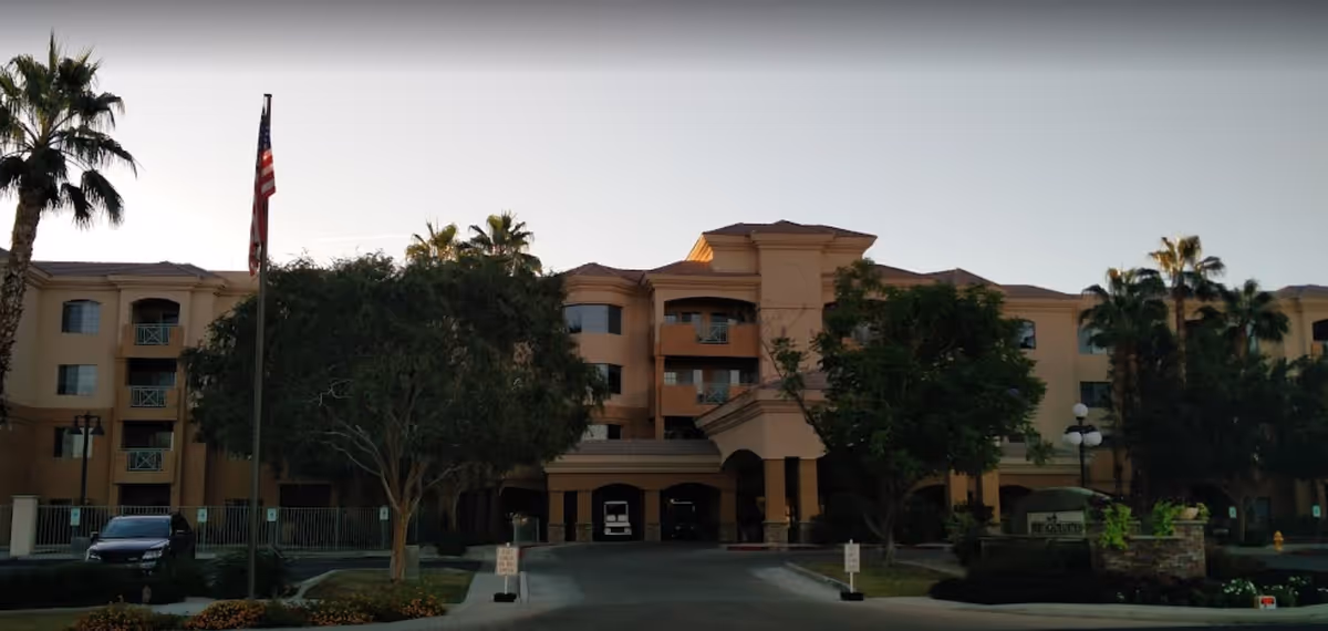Front exterior of a multi-story beige senior living building with palm trees, a flagpole, parked cars, and a covered main entrance.