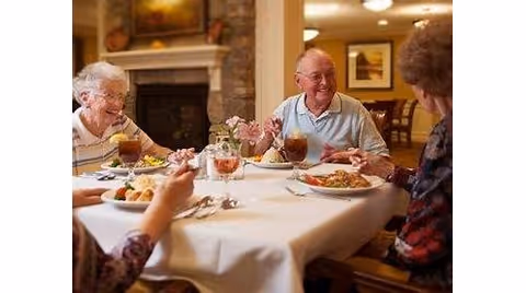 A group of elderly people sitting around a dining table enjoying a meal together in a warm, well-lit room with a fireplace in the background.