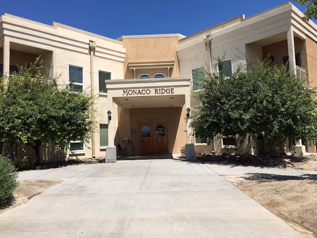 Entrance of a two-story stucco building with a 'MONACO RIDGE' sign above double doors and trees flanking the driveway.