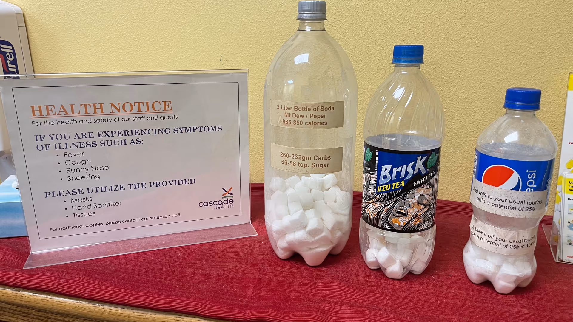 Health notice sign and three soda bottles filled with sugar cubes on a red counter.