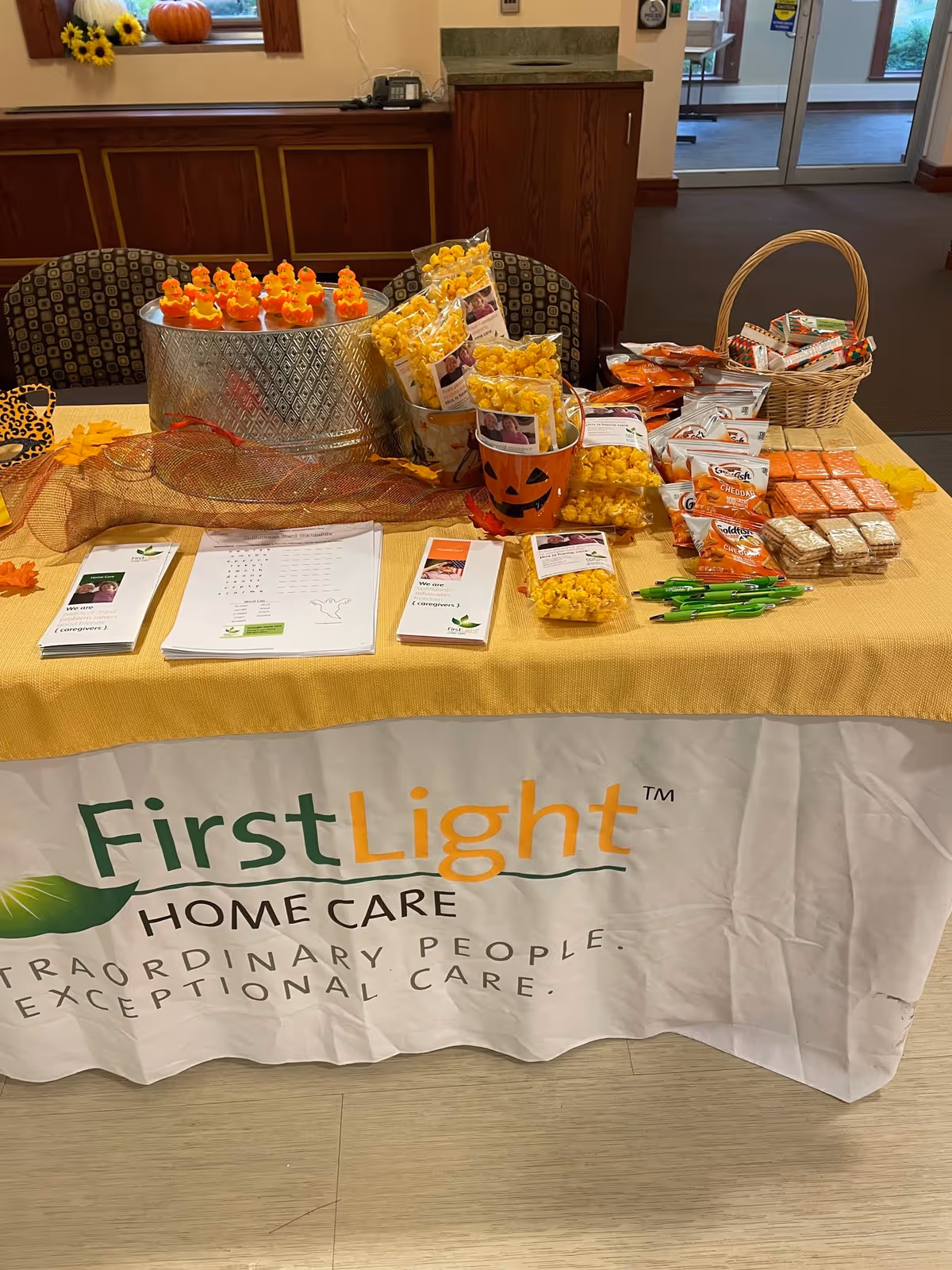Table with FirstLight Home Care banner and snacks inside a facility lobby.