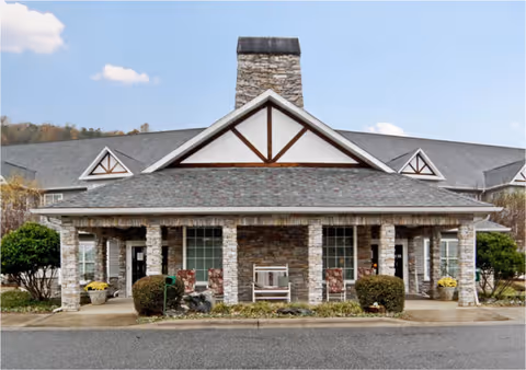 Front entrance of a stone-faced senior living building with a covered porch, columns, rocking chairs, and a central chimney.