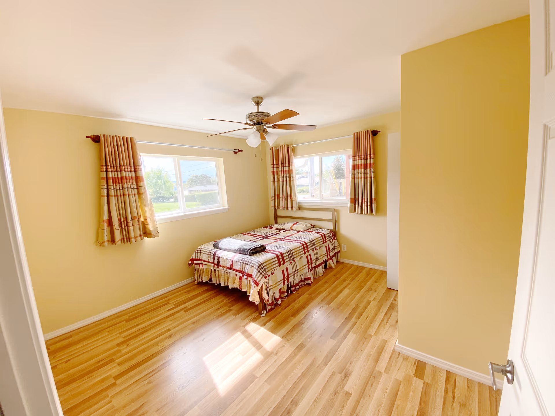 Bright yellow bedroom with a bed, plaid bedding, two windows, and a ceiling fan.
