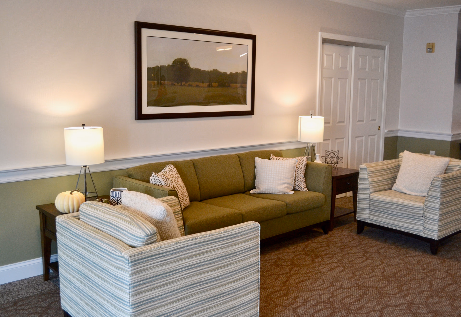 A cozy living room area with a green sofa and two striped armchairs, each with cushions. There are two side tables with lamps on either side of the sofa. A framed landscape picture hangs on the wall above the sofa. The room has a carpeted floor and light-colored walls with a green lower half and white trim.