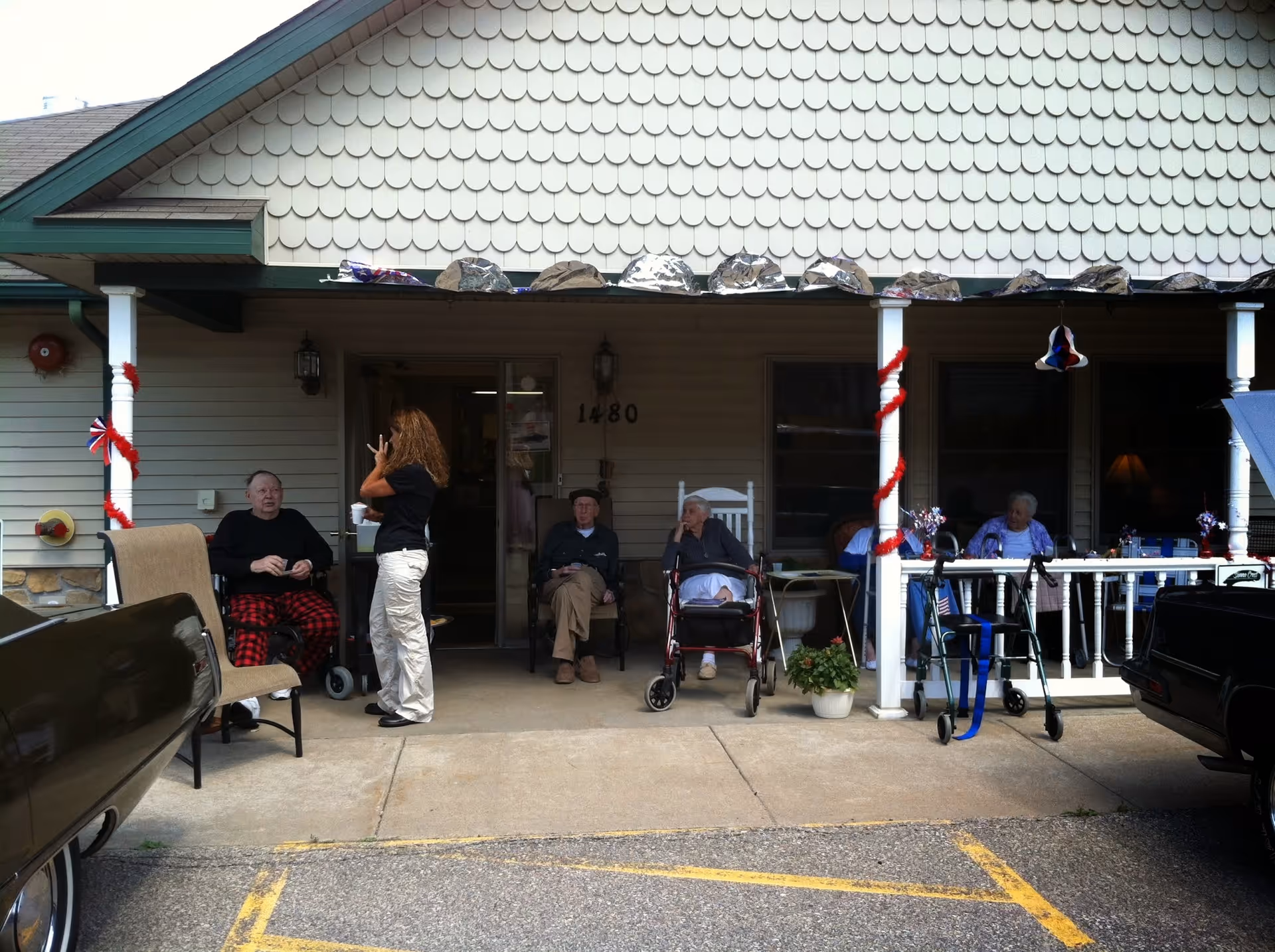 Several elderly people sitting outside on a porch of a building with a woman standing and talking to one of them. The porch is decorated with red, white, and blue ribbons and small patriotic decorations. There are walkers and chairs around, and the building has a scalloped shingle exterior with the number 1480 visible near the door.