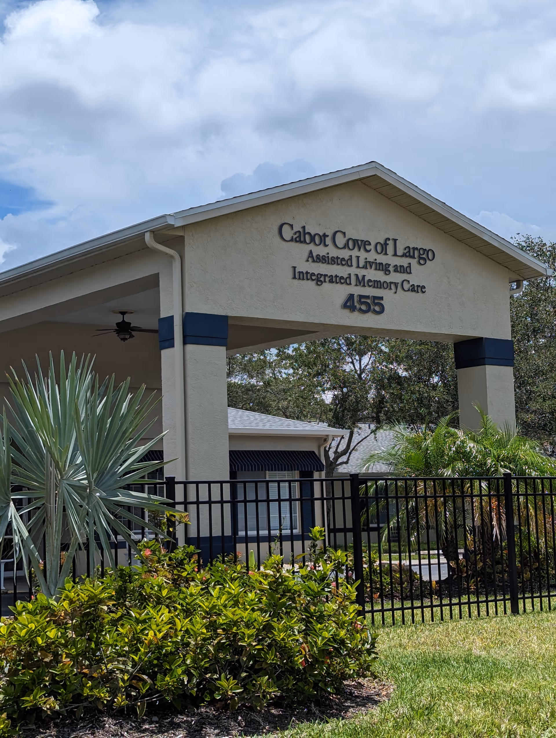 Entrance canopy and sign reading 'Cabot Cove of Largo Assisted Living and Integrated Memory Care 455' with landscaping and a black fence in front.