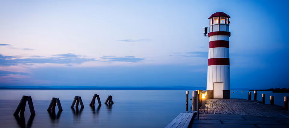A striped lighthouse sits on a wooden pier extending over calm blue water at dusk.