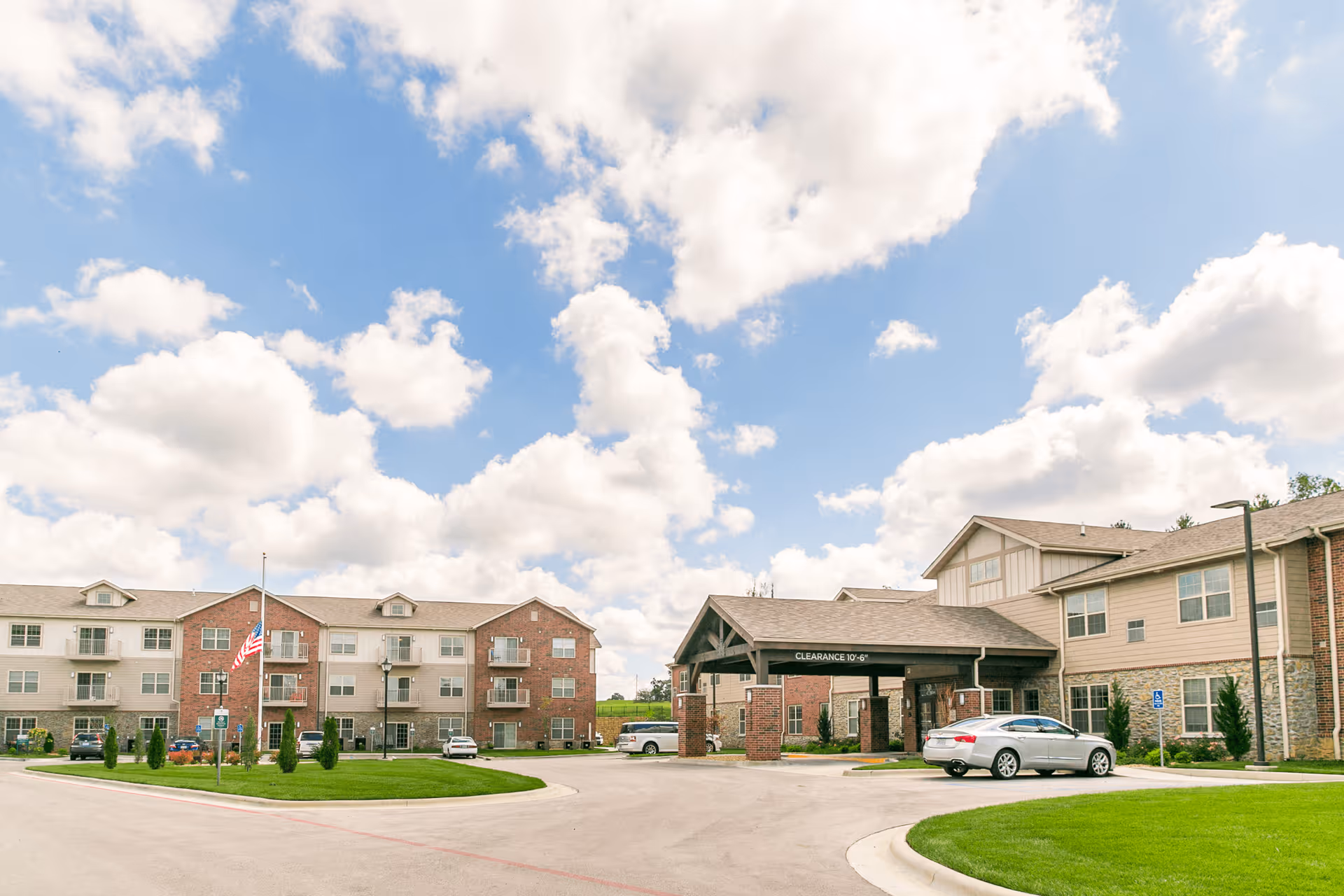 Exterior view of a senior living facility with multiple connected buildings, a covered entrance with a clearance sign, a driveway, parked cars, a flagpole with an American flag, and a well-maintained lawn under a partly cloudy sky.