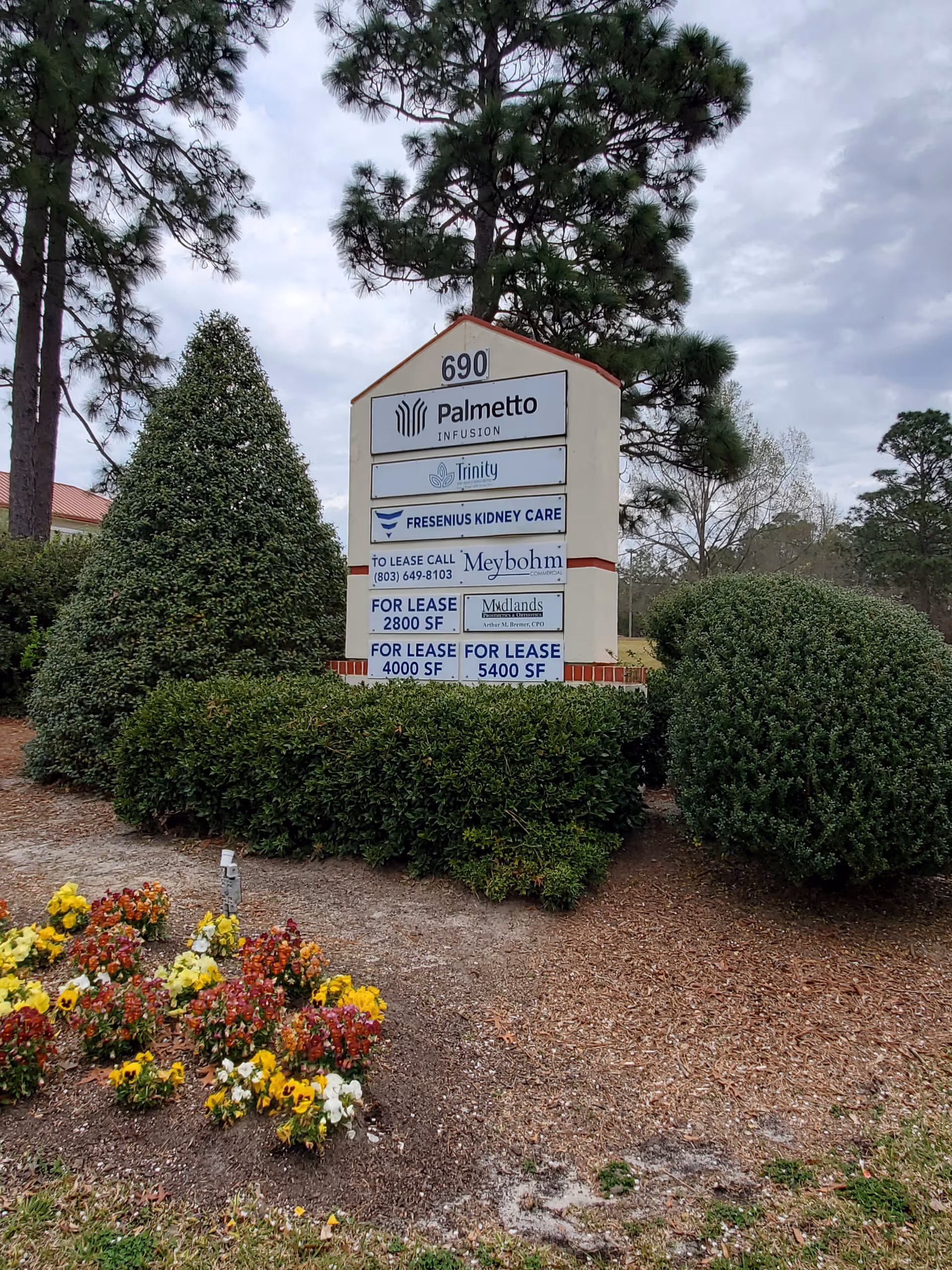 Outdoor sign with multiple business names amid trimmed bushes and colorful flowers