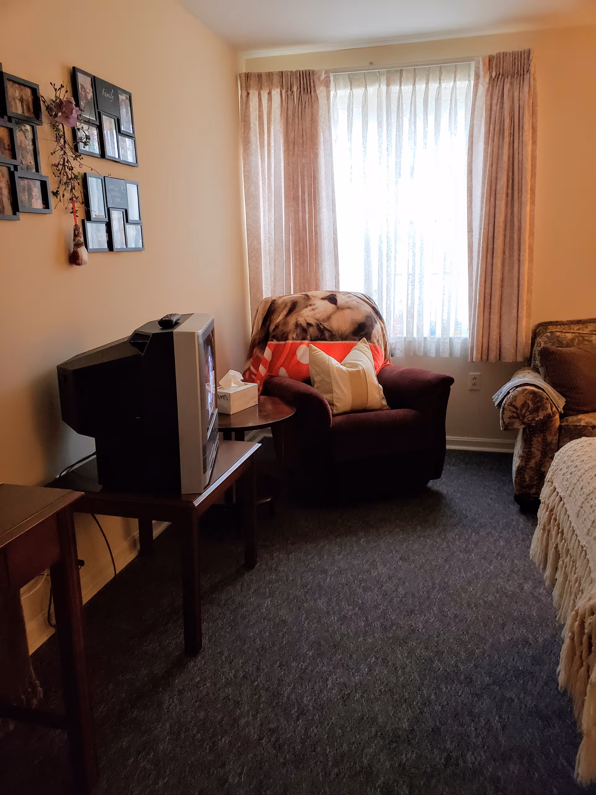 Cozy living area in a senior unit with chair, TV, curtains, and decorative wall frames.