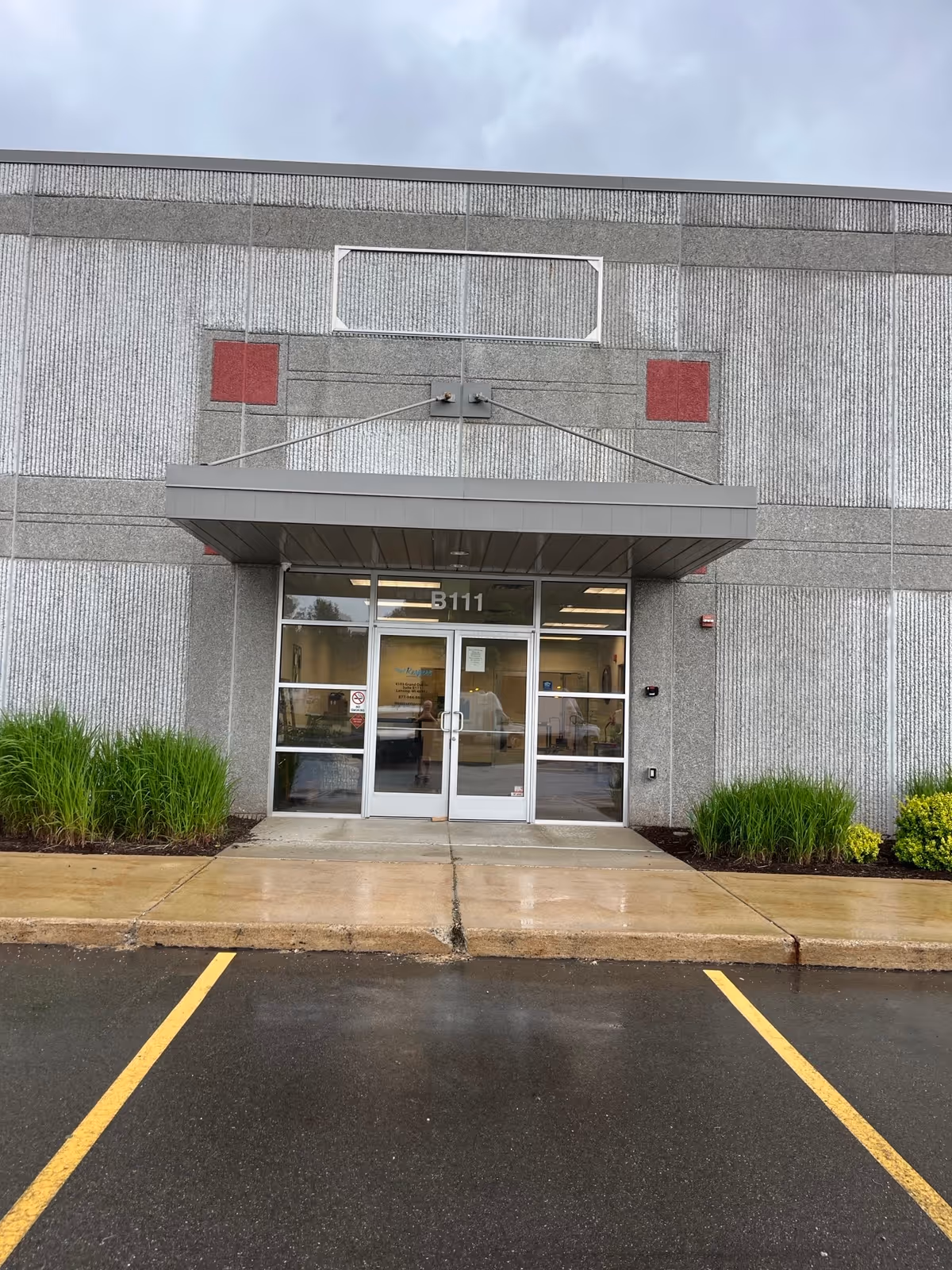 Grey building entrance with glass doors and metal awning, wet parking lot in front.