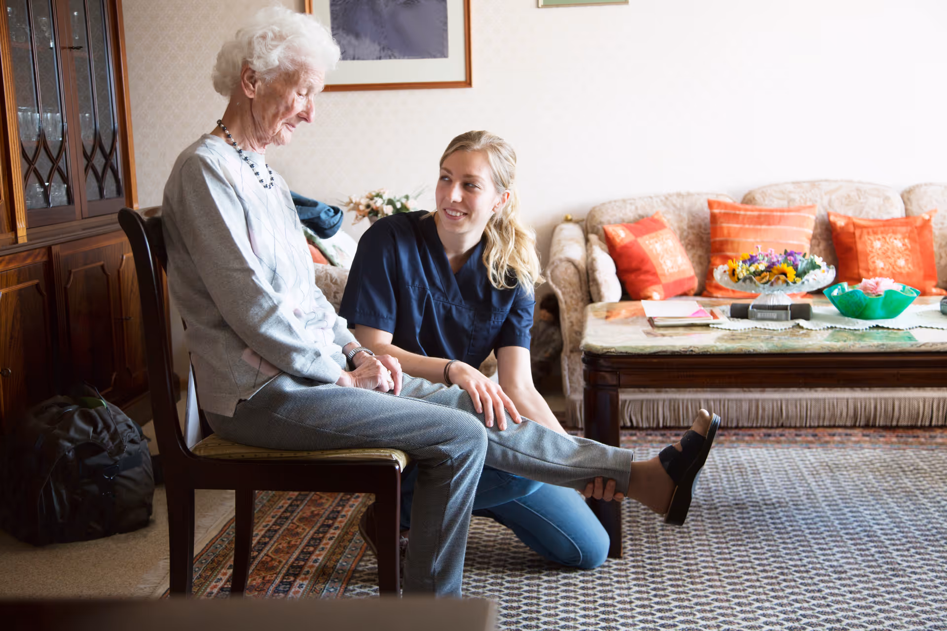 Elderly woman sits on a chair while a caregiver kneels beside her in a living room.