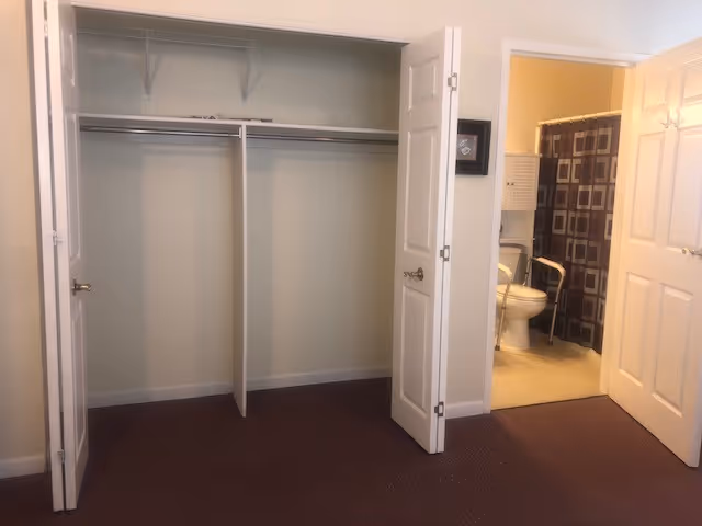 Open closet with hanging rod and shelf beside doorway to bathroom in a bedroom.