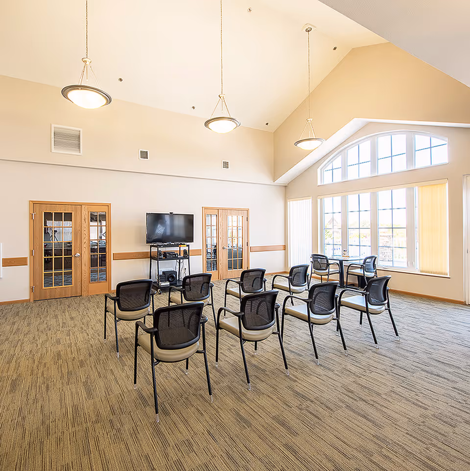A bright, spacious room with high vaulted ceilings and large windows letting in natural light. The room has a carpeted floor and is furnished with several black mesh chairs arranged in rows facing a flat-screen TV mounted on a stand. There are two wooden doors with glass panels on one wall and a small table with two chairs near the windows.