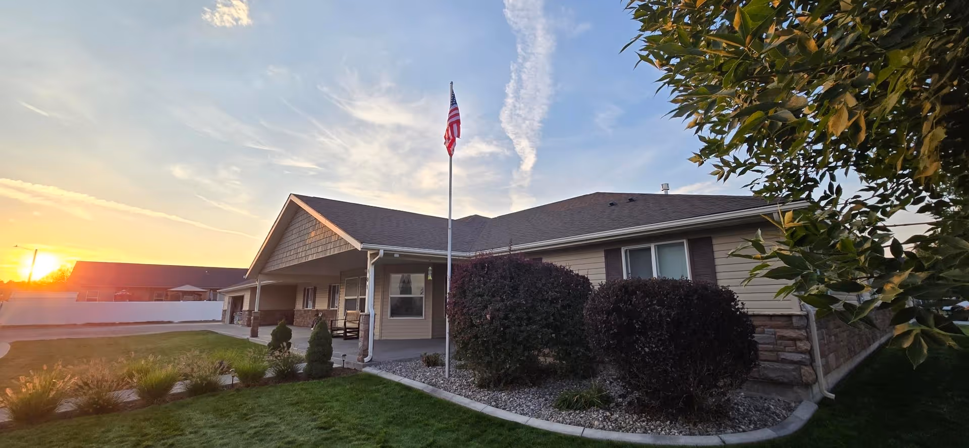Single-story Ashley Manor Memory Care exterior with flagpole, sunset, and manicured shrubs.