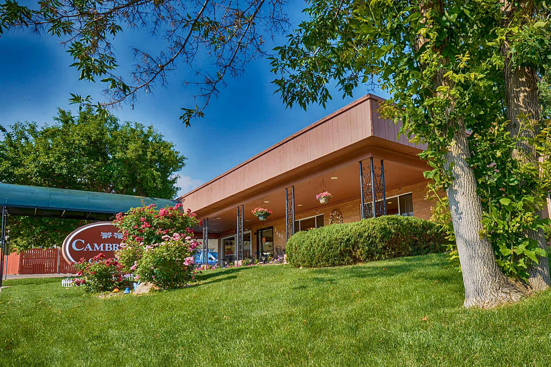 Front exterior of the Cambridge Care Center building with a green lawn, flowering bushes, hanging baskets, and a covered entrance.