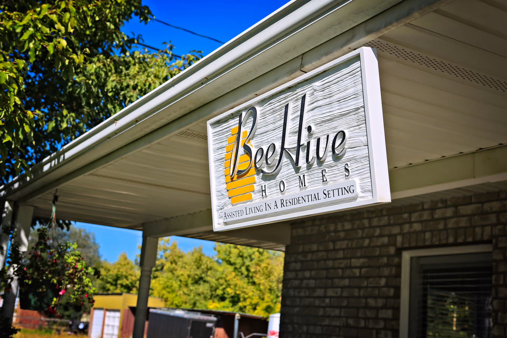 Hanging wooden sign reading 'Beehive Homes' on a porch overhang of a brick building with trees in the background.