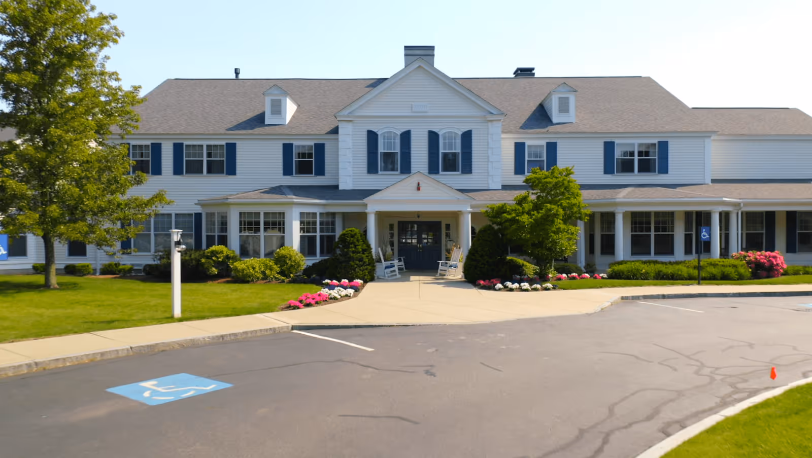 Front exterior view of a two-story white building with blue shutters, a gray roof, and a covered entrance with two rocking chairs. The building is surrounded by green grass, trees, and colorful flower beds. There is a paved driveway with a handicap parking space in front.