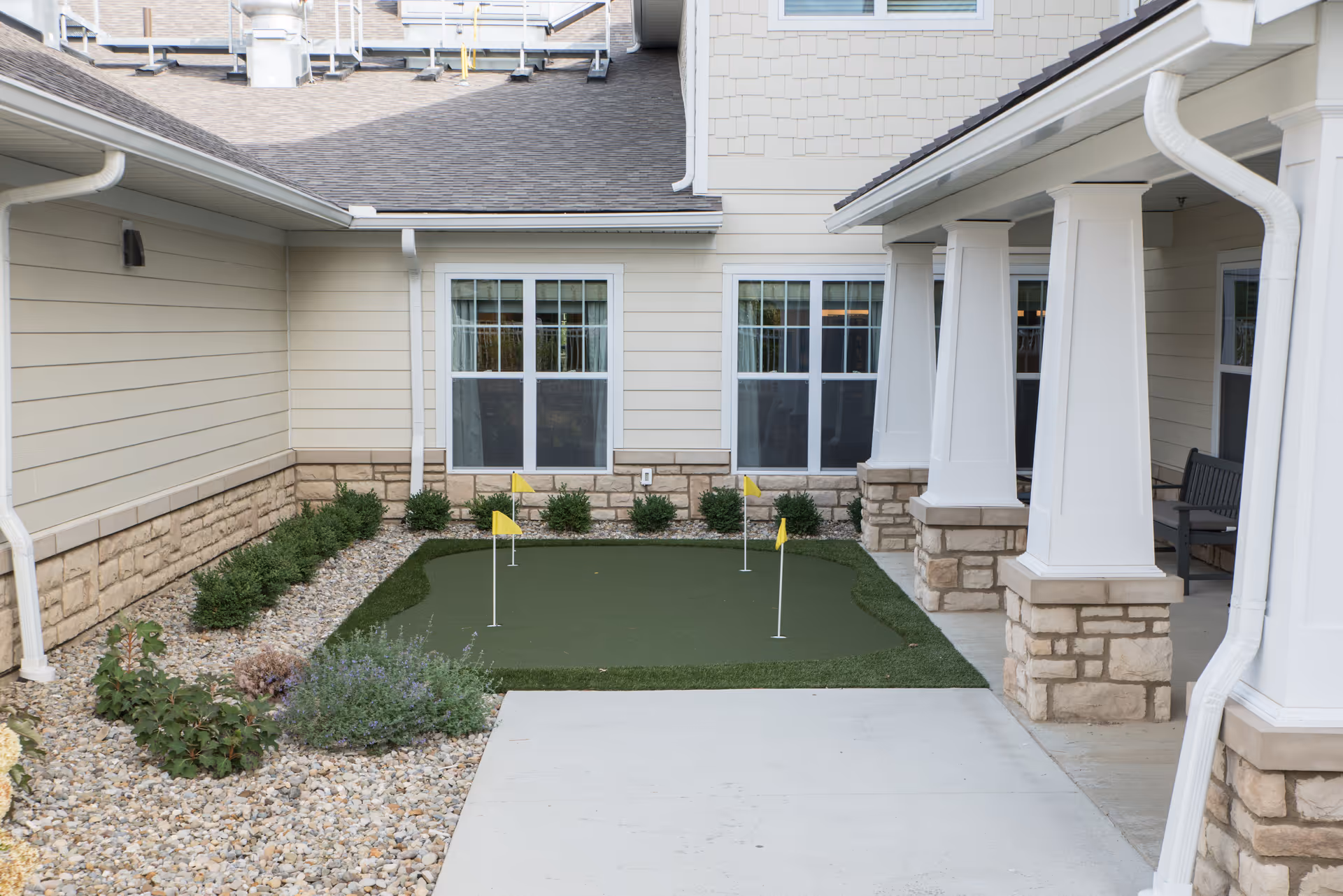 A small courtyard putting green with three yellow flags in front of a beige senior living building with columns, windows, and a bench.