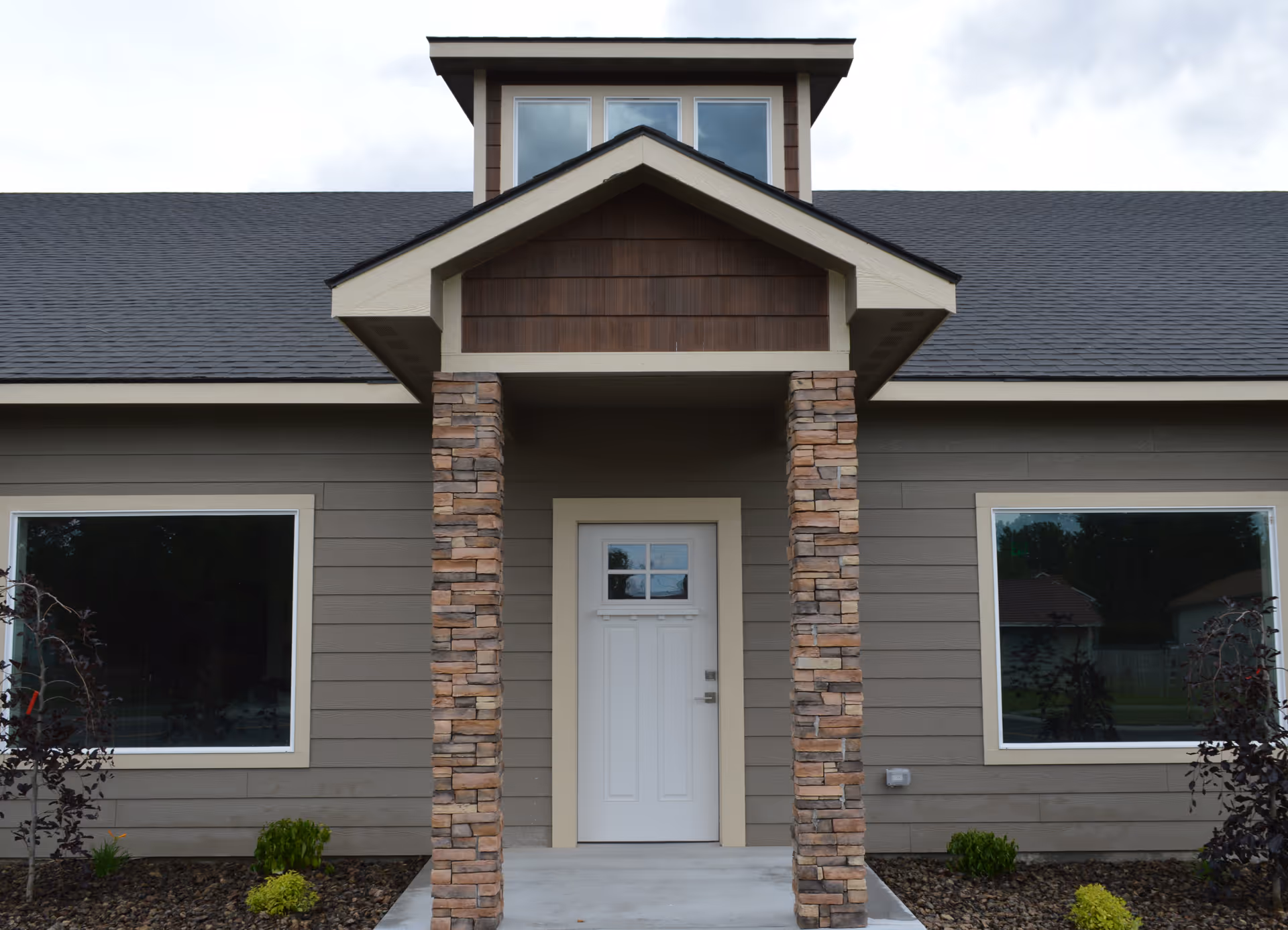 Front exterior view of a single-story building with a white door centered between two large windows. The entrance is framed by two stone pillars supporting a small gabled roof. The building has gray siding and a dark shingled roof with a small dormer window above the entrance.