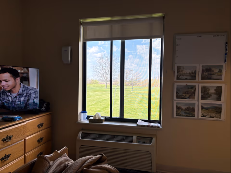 Sunlit window with blinds, an air conditioner, dresser with TV, and a bed in a bedroom.