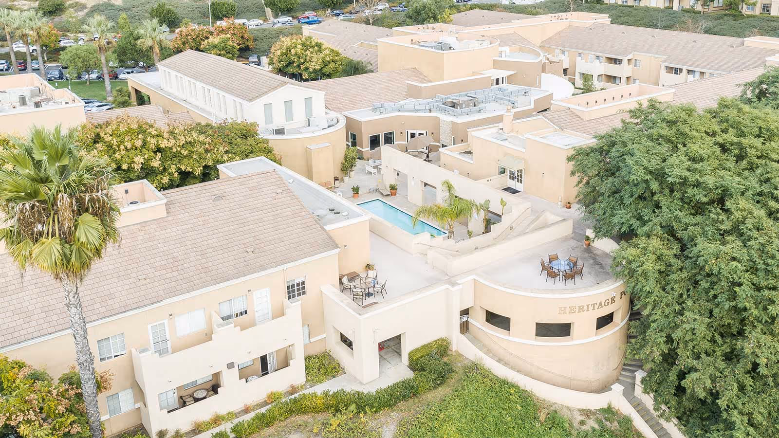 Aerial view of Heritage Pointe senior living facility showing beige buildings with multiple rooftops, outdoor seating areas, a small rectangular pool, surrounded by trees and greenery.
