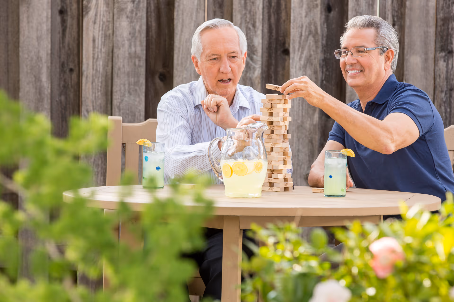 Two seniors sit outdoors at a round table, playing Jenga with lemonade.