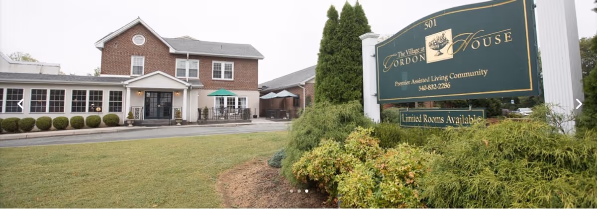 Exterior front of The Village at Gordon House assisted living building with brick façade, entrance, patio and a large green sign in landscaped lawn.