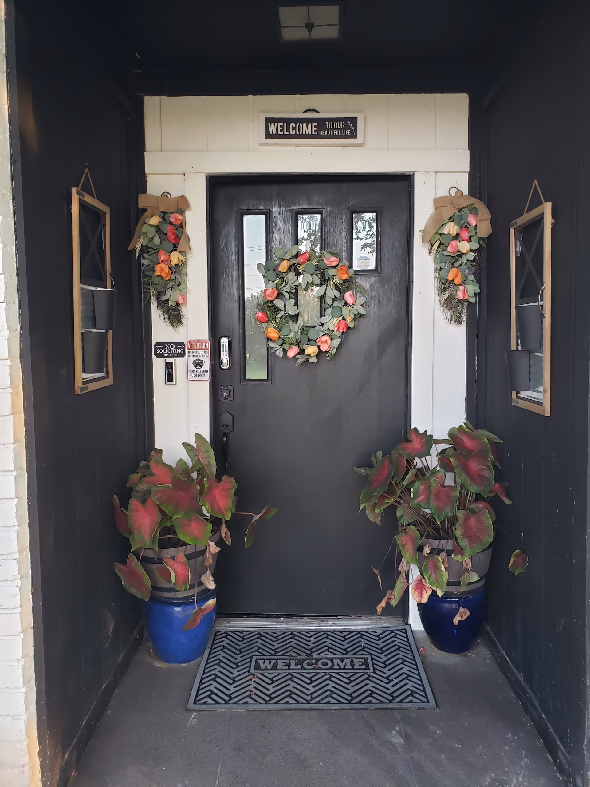 Black front door framed by wreaths, potted plants, and a welcome mat.
