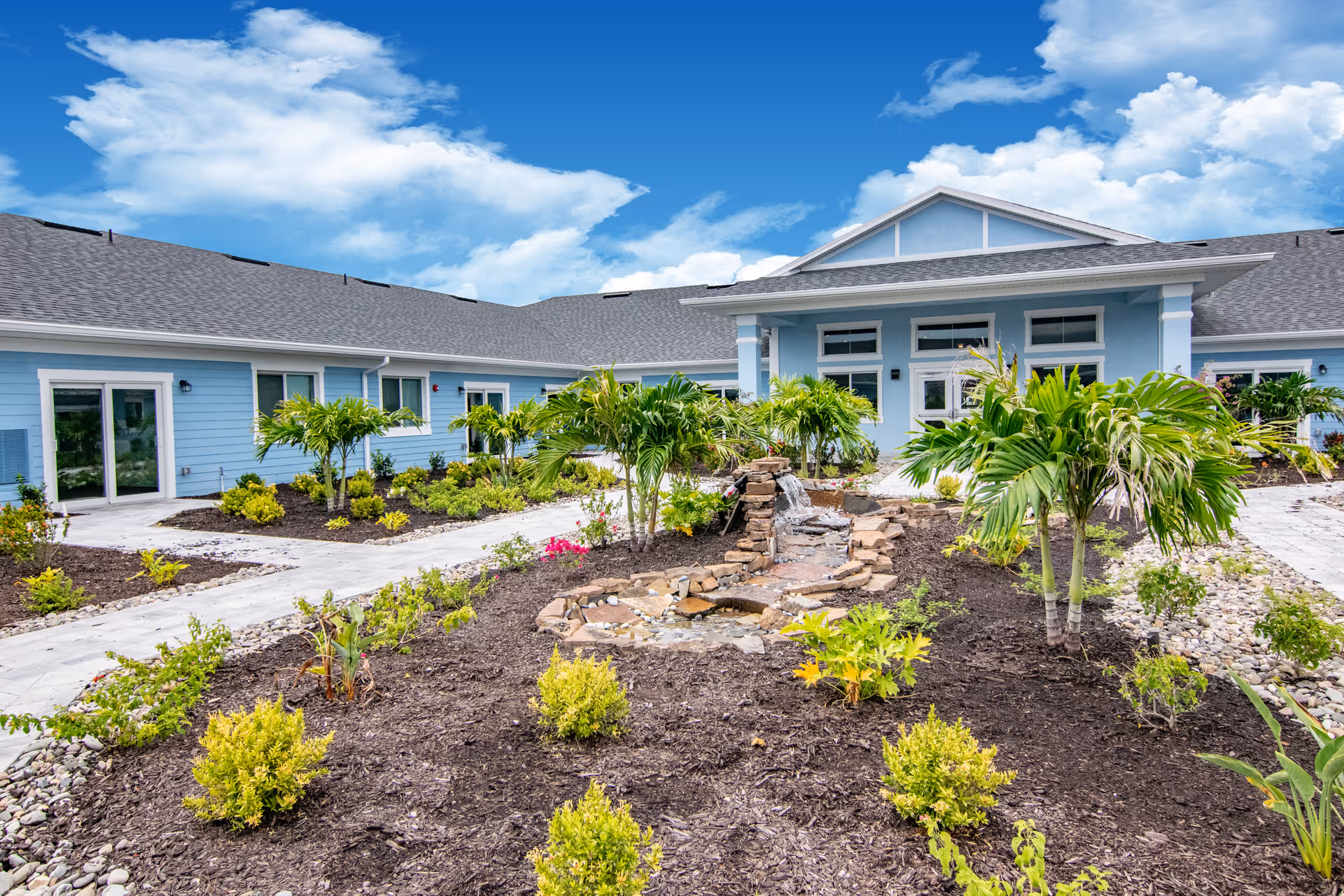 Blue single-story senior living building with landscaped courtyard and palm trees.
