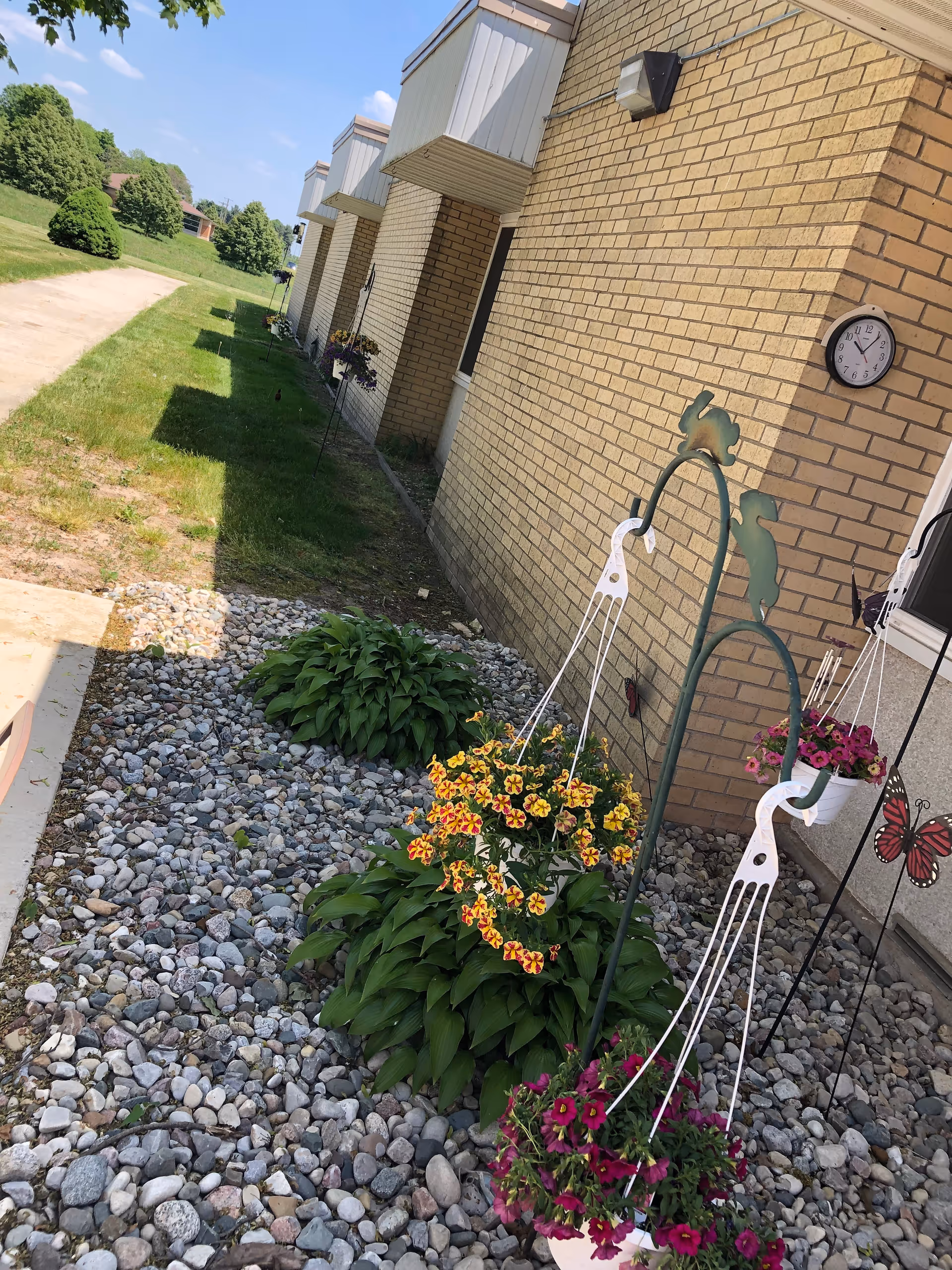 Outdoor view of a brick building with a clock mounted on the wall showing 12:15. In front of the building, there are hanging flower pots with colorful flowers and green plants surrounded by small rocks. A grassy area and a paved path are visible to the left, with trees and a clear blue sky in the background.