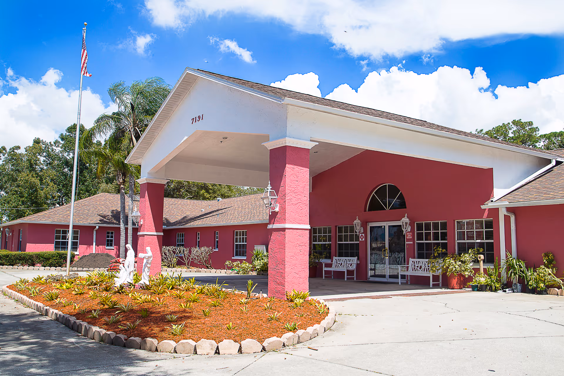 Front entrance of a pink assisted living building with a porte-cochère, landscaped planter and an American flag.