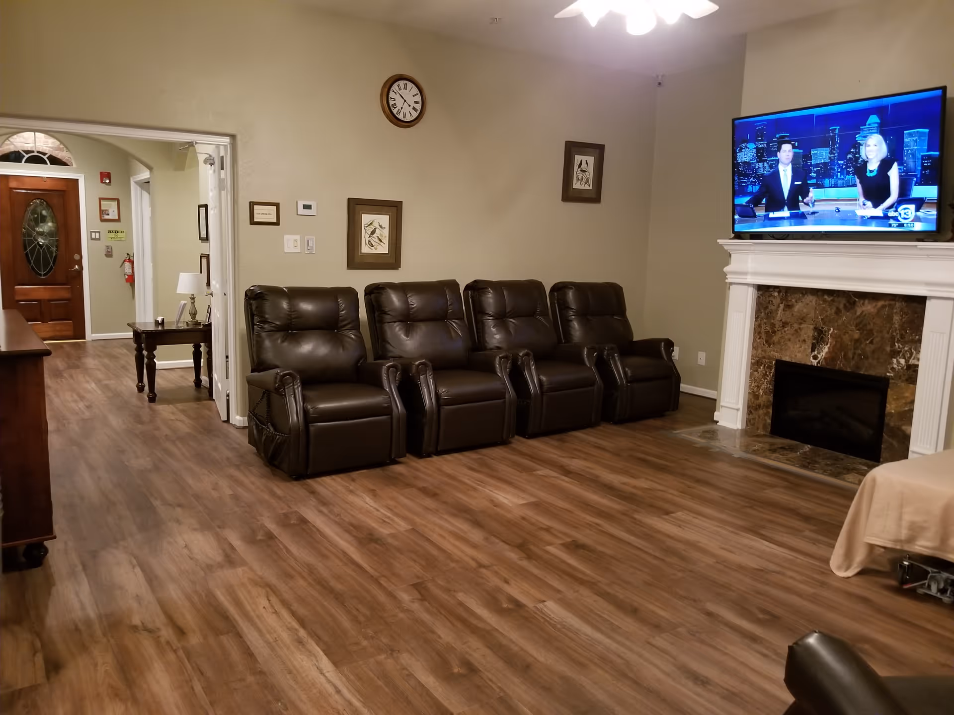 A cozy living room with four dark brown leather recliner chairs lined up against a beige wall. Above the chairs are two framed pictures and a round wall clock. A flat-screen TV is mounted above a white fireplace with a brown marble surround, displaying a news broadcast. The room has wood flooring and a ceiling fan with lights. In the background, there is a hallway leading to a wooden front door with glass panels and a small table with a lamp.