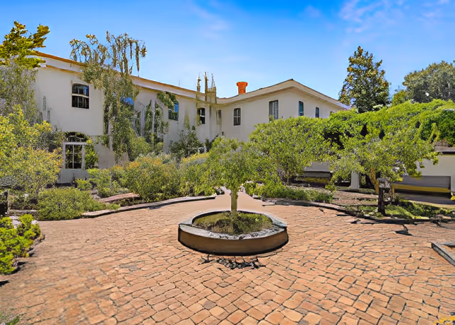 Brick courtyard with a circular planter and trimmed trees in front of a two-story white building under a blue sky.