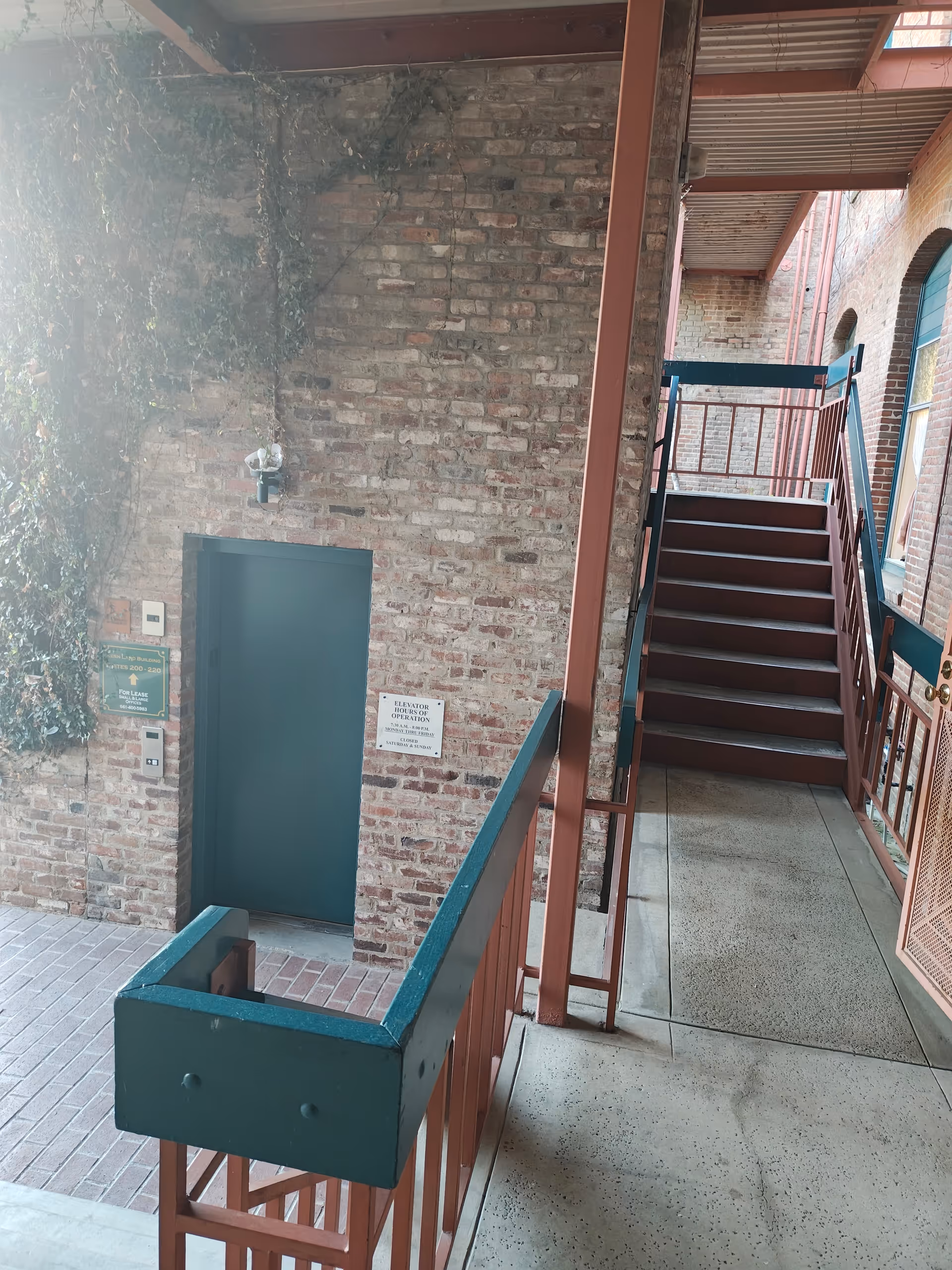 Brick-walled interior stairwell with teal railings, a green door, and elevated walkway.