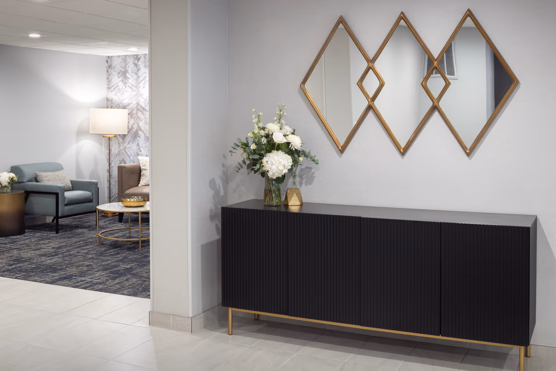 Modern lobby area with a black sideboard, geometric mirrors, a flower arrangement, and seating.