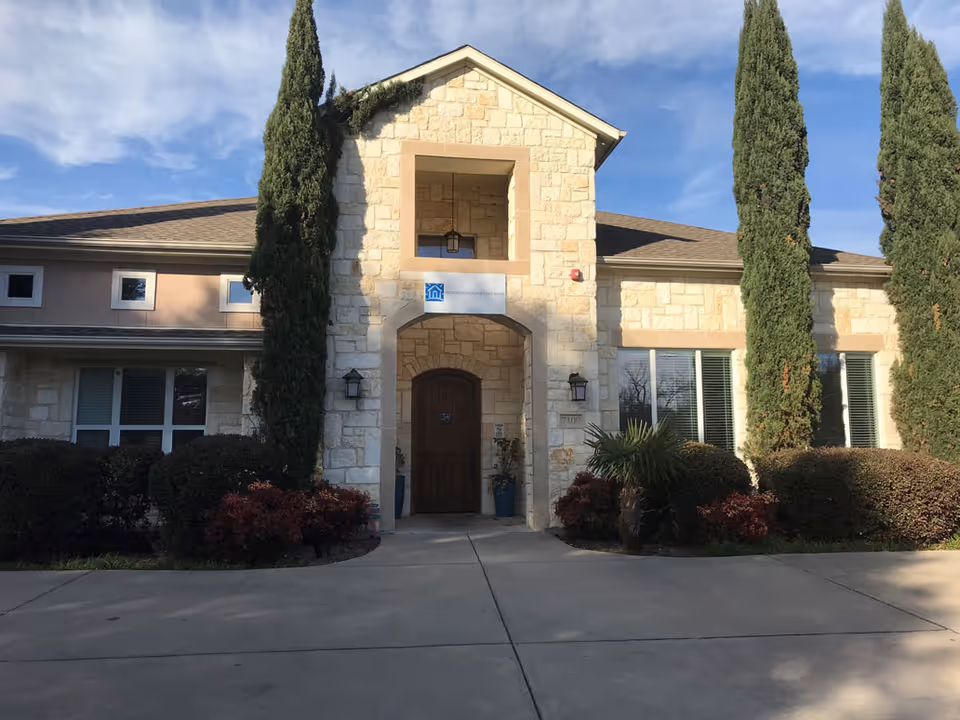 Stone-faced senior living facility entrance with archway, shrubbery, and tall cypress trees.