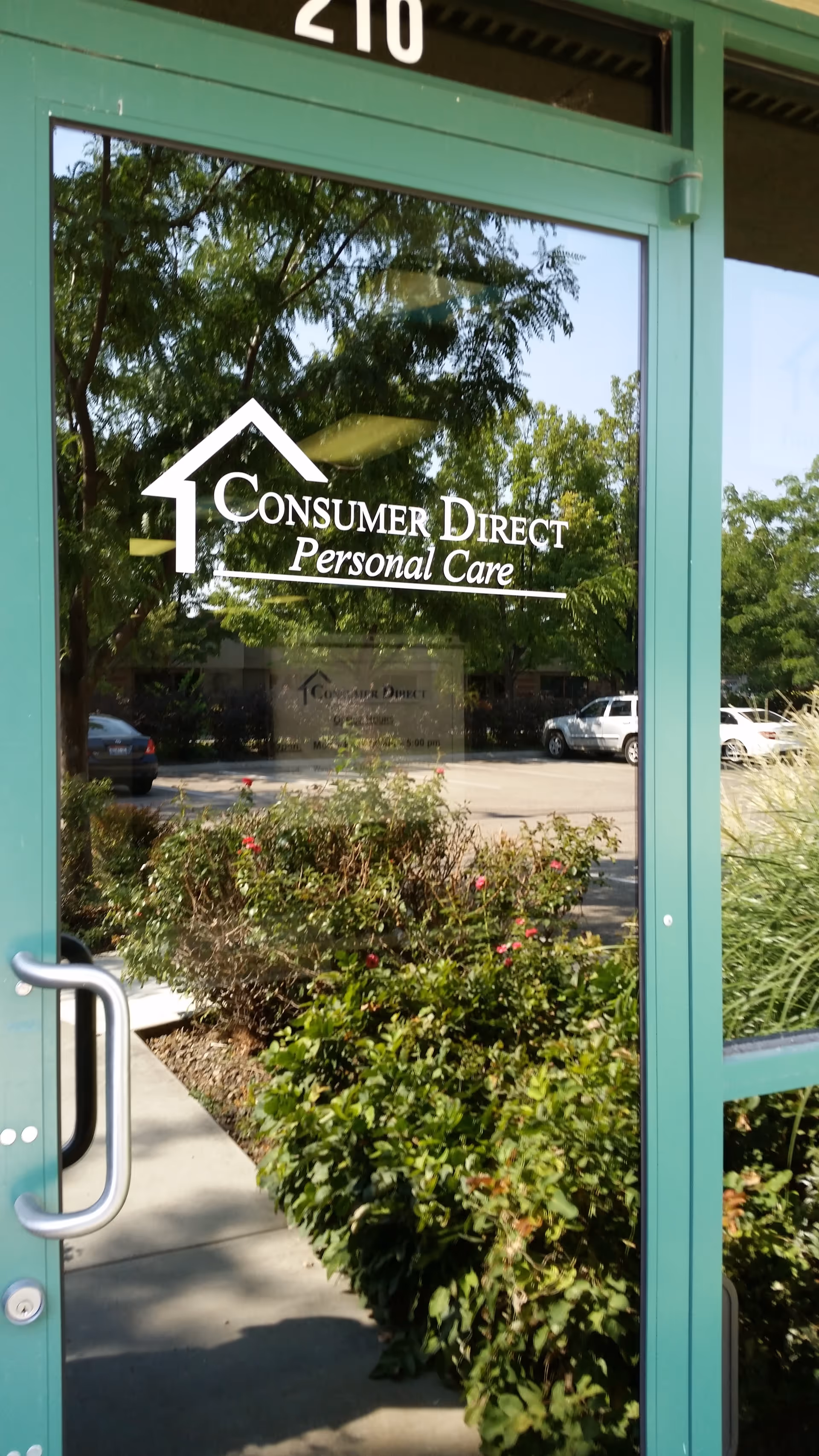 Green-framed glass entrance door displays Consumer Direct Personal Care logo with landscaping outside.