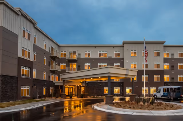 Three-story senior living building with a covered entrance, driveway, flagpole, and lit windows at dusk.