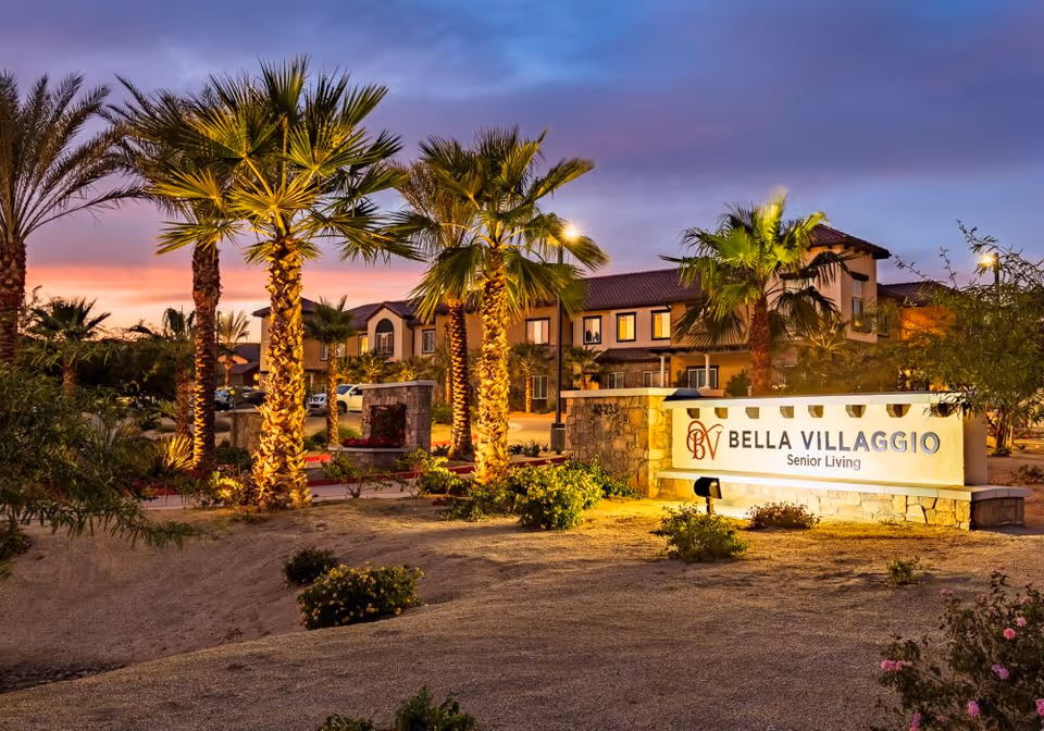 Exterior view of Bella Villaggio Senior Living with palm trees and a lit entrance sign.