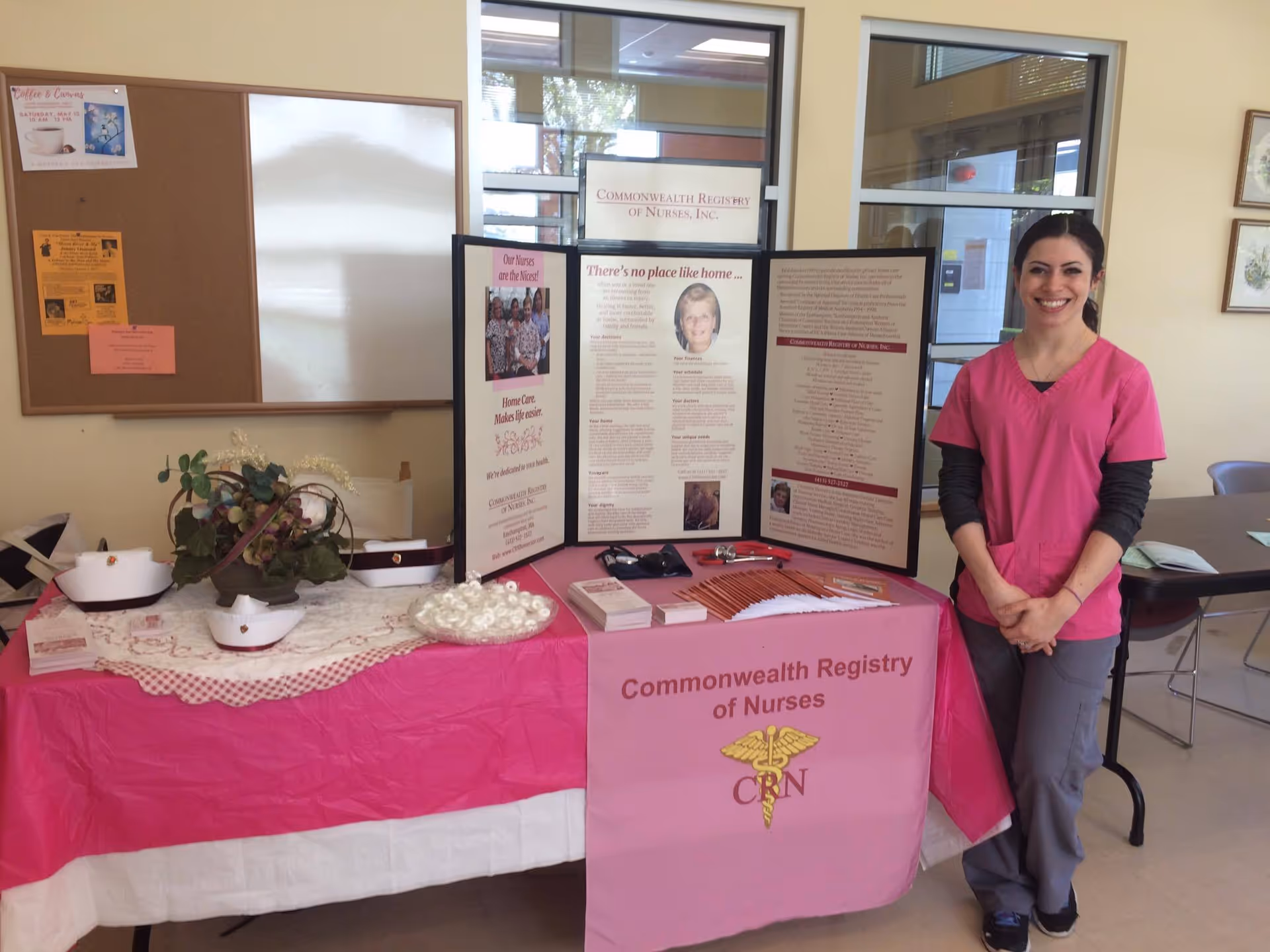 A staff member in pink scrubs stands beside a nursing display table indoors.