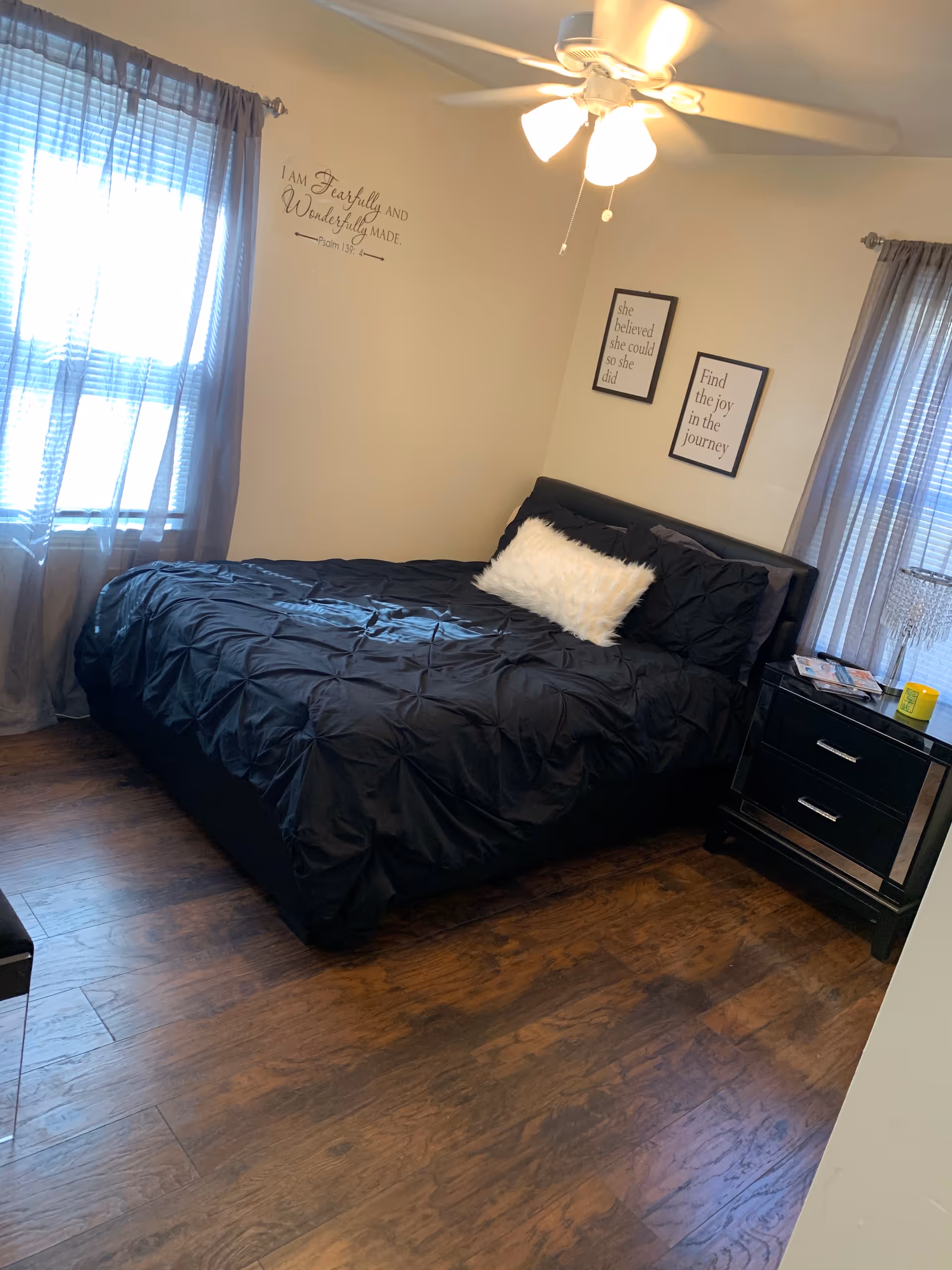 Cozy bedroom with a dark comforter, white fluffy pillow, curtains, nightstand, and wall quotes.