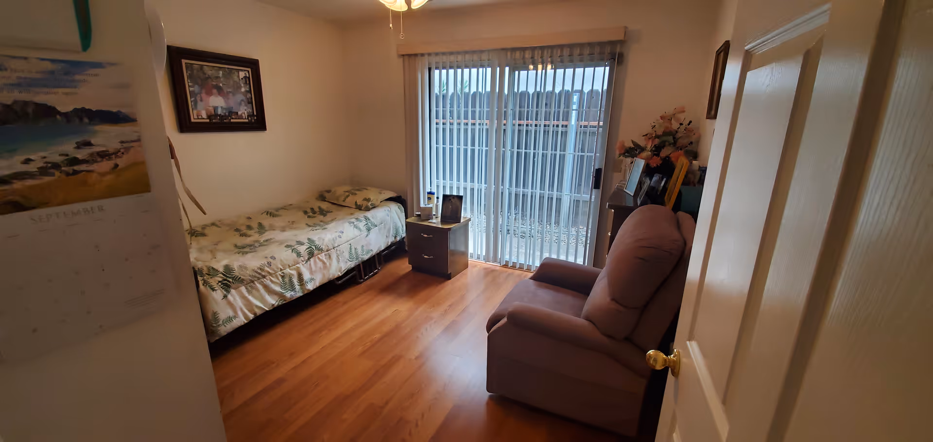 Small bedroom in a senior living unit featuring a bed, recliner, and sliding glass door.