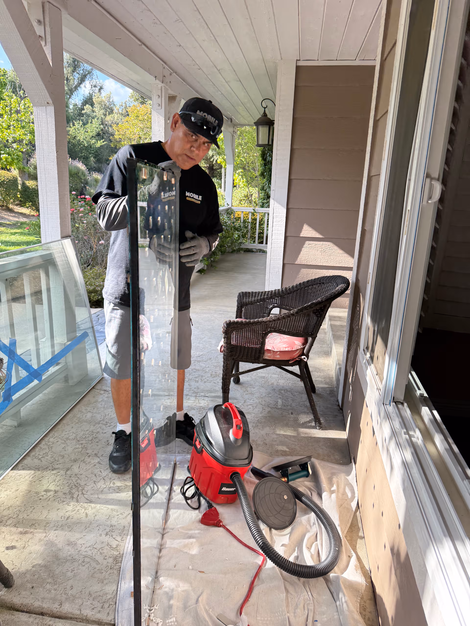 Man wearing gloves cleans a glass pane on a covered porch, with a red vacuum nearby.